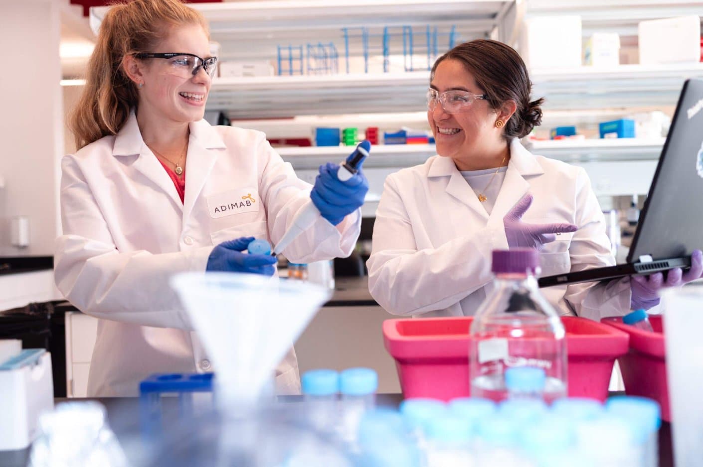 Two scientists in a lab working with pipettes and a laptop.